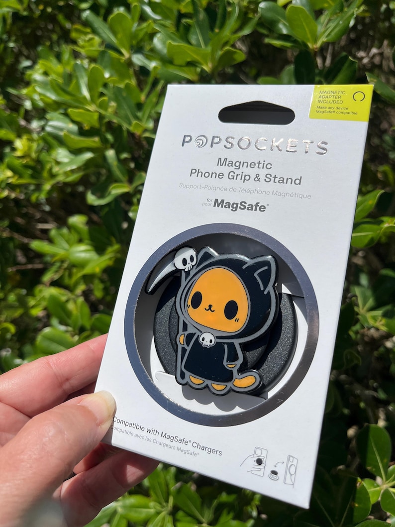 May include: A PopSockets magnetic phone grip and stand featuring a cartoon Grim Reaper cat design. The black and orange character holds a scythe and wears a hooded robe. The packaging includes the text "Magnetic Phone Grip & Stand" and "Compatible with MagSafe Chargers."