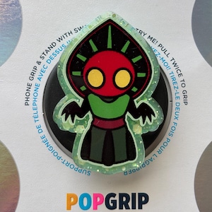 May include: A PopGrip phone accessory featuring a cartoon character design. The character has a red face with yellow eyes, a green body, and a black and green outfit. The accessory is black with text around the edge.