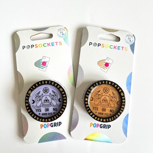 May include: Two phone grips with a crystal ball and hands design. The phone grips are black with a gold outline and text. One phone grip has a purple background and the other has a wood background. The text on the phone grips reads "YES OR NO + GOOD BYE".