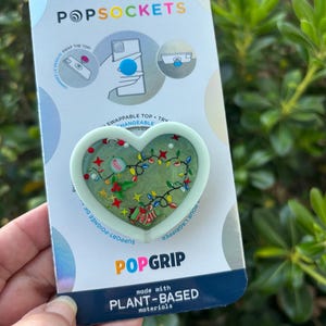 May include: A PopSockets PopGrip with a heart-shaped design, filled with festive Christmas-themed decorations. The PopGrip is mint green and features the text "POPGRIP" and "made with PLANT-BASED materials" on the packaging.