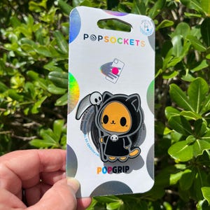May include: A PopSocket PopGrip featuring a cartoon cat dressed as the Grim Reaper, with a scythe and skull. The PopSocket is on a white and holographic backing with the PopSockets logo and the words "PopGrip".