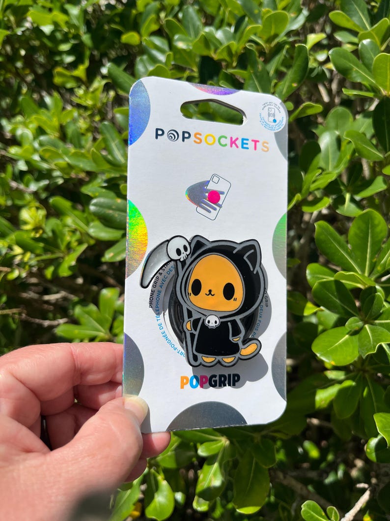 May include: A PopSocket PopGrip featuring a cartoon cat dressed as the Grim Reaper, with a scythe and skull. The PopSocket is on a white and holographic backing with the PopSockets logo and the words "PopGrip".