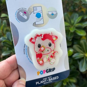 May include: A PopSockets PopGrip phone grip featuring a white cow design with red accents and strawberries. The PopGrip is made with plant-based materials and is attached to a white and blue backing with the PopSockets logo.