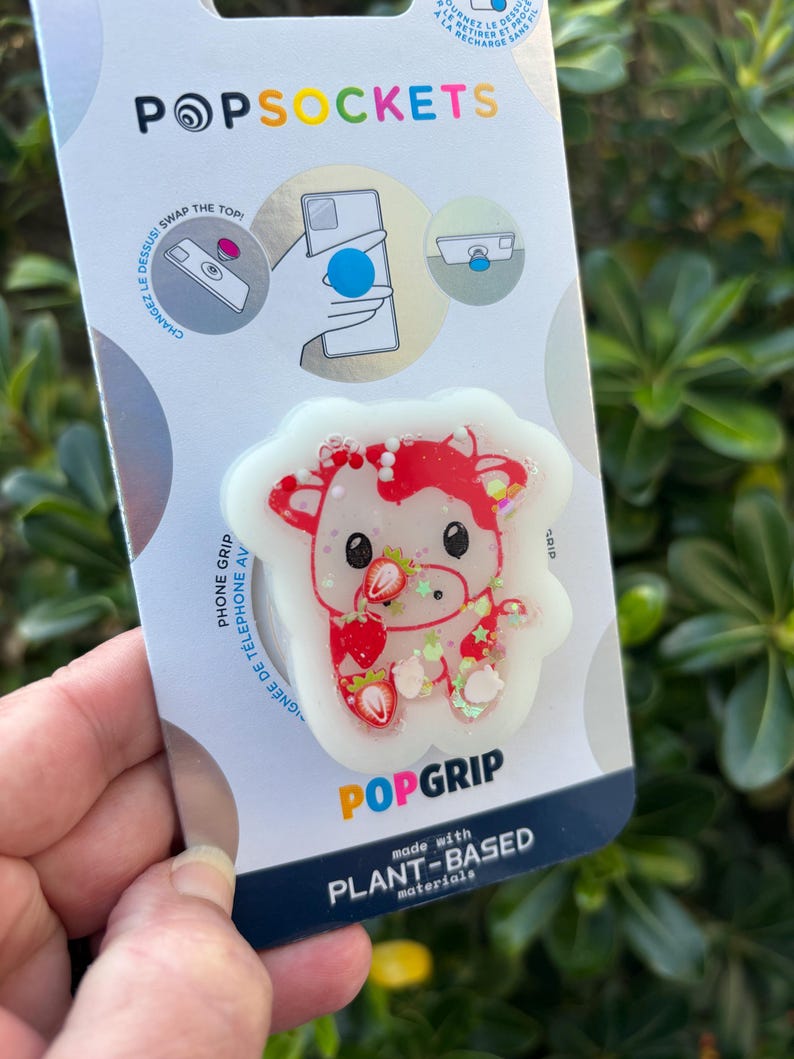 May include: A PopSockets PopGrip phone grip featuring a white cow design with red accents and strawberries. The PopGrip is made with plant-based materials and is attached to a white and blue backing with the PopSockets logo.