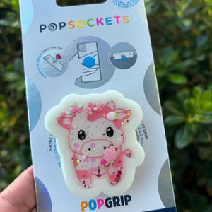 May include: A PopSockets PopGrip featuring a pink and white cow design with glitter accents. The packaging includes the PopSockets logo and text that reads "made with PLANT-BASED materials."
