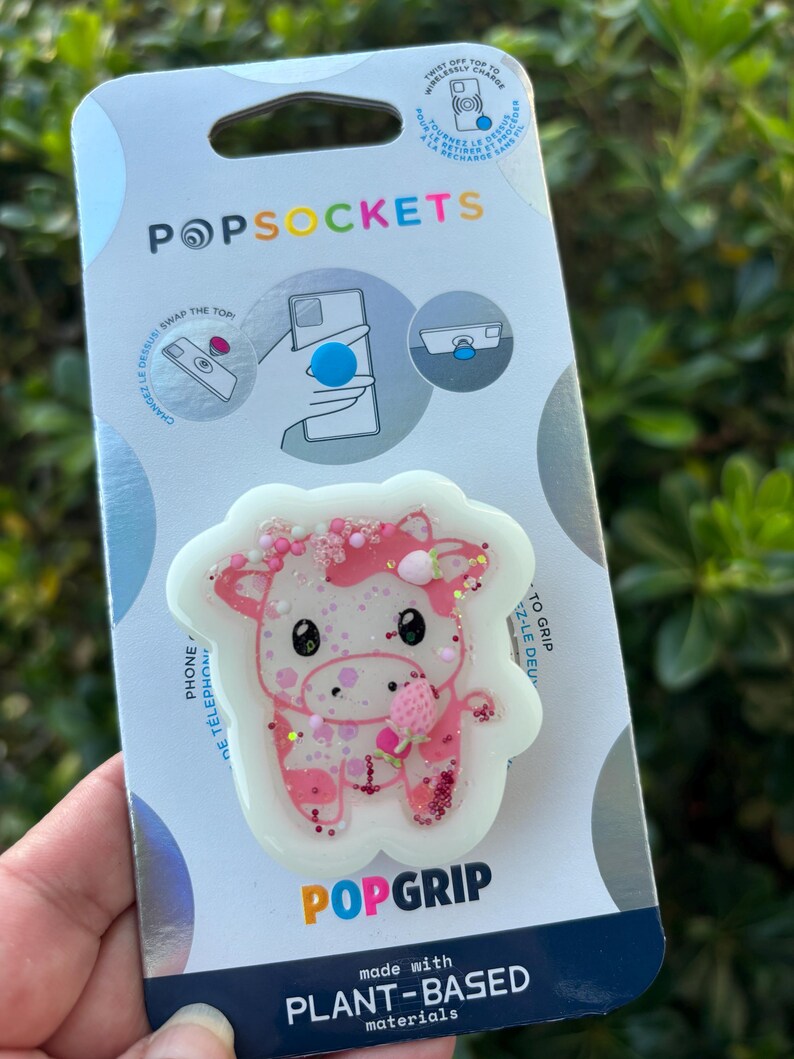 May include: A PopSockets PopGrip featuring a pink and white cow design with glitter accents. The packaging includes the PopSockets logo and text that reads "made with PLANT-BASED materials."