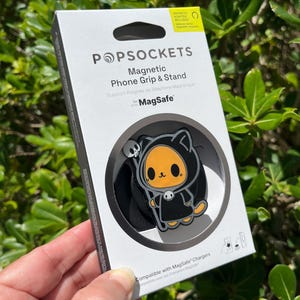 May include: A white box with a black and orange cartoon character PopSocket phone grip and stand. The box reads "POPSOCKETS Magnetic Phone Grip & Stand for MagSafe". The PopSocket has a black background.