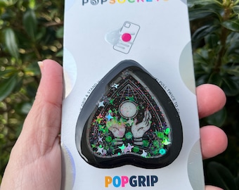 May include: A black heart-shaped phone grip with a green and black glitter design. The grip features a white circle with a black outline and two white hands with black outlines reaching towards the circle. The grip is labeled "POPGRIP" and "POPSOCKETS" on the packaging.