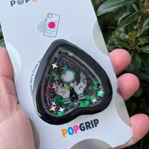Pu&ograve; includere: Un'impugnatura per telefono nera a forma di cuore con un design glitterato che presenta un paio di mani che tengono una sfera di cristallo e stelle. L'impugnatura &egrave; attaccata a una carta bianca con il testo "POPSOCKETS" e "POPGRIP".