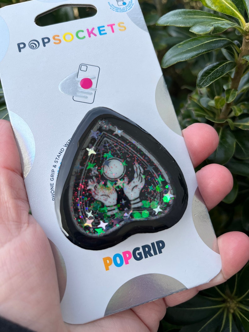 Pu&ograve; includere: Un'impugnatura per telefono nera a forma di cuore con un design glitterato che presenta un paio di mani che tengono una sfera di cristallo e stelle. L'impugnatura &egrave; attaccata a una carta bianca con il testo "POPSOCKETS" e "POPGRIP".