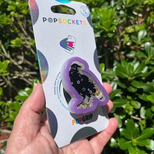 May include: A PopSockets PopGrip featuring a purple owl-shaped design with a black starry background, gold accents, and the text "PopGrip" in orange. The PopSockets packaging is visible.