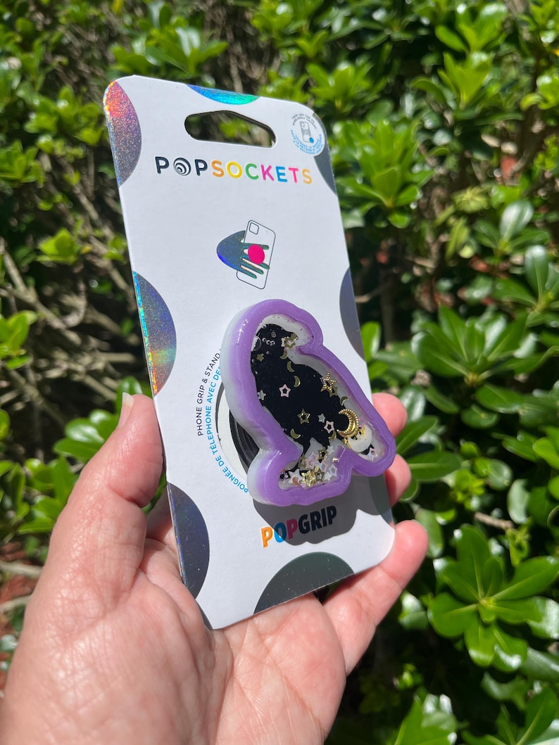 May include: A PopSockets PopGrip featuring a purple owl-shaped design with a black starry background, gold accents, and the text "PopGrip" in orange. The PopSockets packaging is visible.