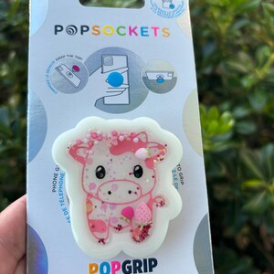 May include: A PopSockets PopGrip featuring a pink cow design with glitter accents. The cow has a strawberry and a pink bow. The PopGrip is on a white background and is attached to a PopSockets branded card.