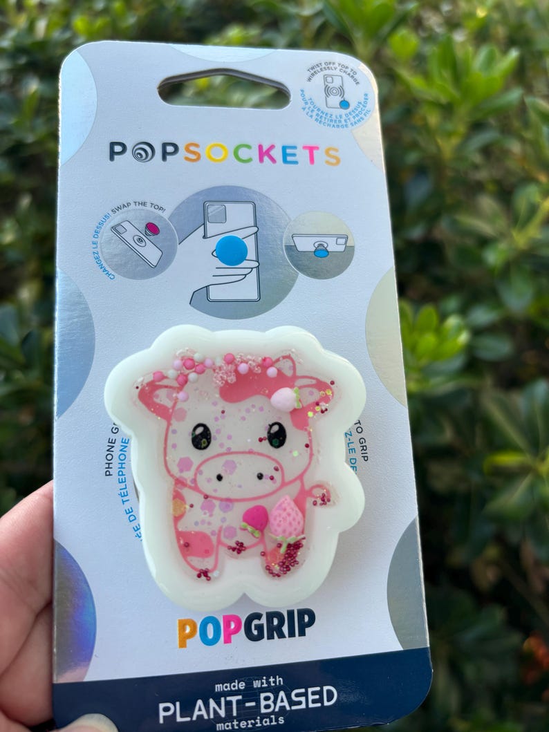 May include: A PopSockets PopGrip featuring a pink cow design with glitter accents. The cow has a strawberry and a pink bow. The PopGrip is on a white background and is attached to a PopSockets branded card.