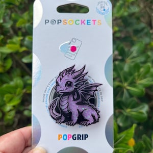 May include: A PopSockets PopGrip featuring a purple dragon design. The dragon has large eyes and detailed wings. The PopGrip is on a silver and white backing with the PopSockets and PopGrip logos.