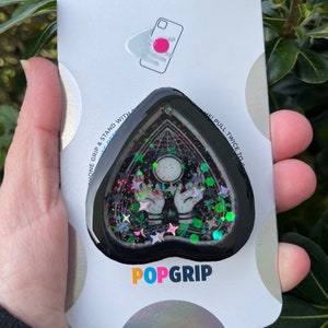 Pu&ograve; includere: Un PopGrip per telefono a forma di cuore nero con un design scintillante che presenta una luna crescente, due mani e stelle. Il testo "POPGRIP" &egrave; stampato sulla confezione.