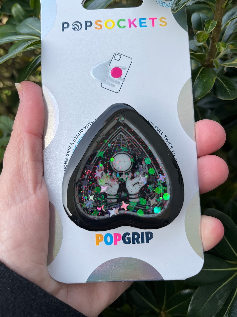 Pu&ograve; includere: Un PopGrip per telefono a forma di cuore nero con un design scintillante che presenta una luna crescente, due mani e stelle. Il testo "POPGRIP" &egrave; stampato sulla confezione.