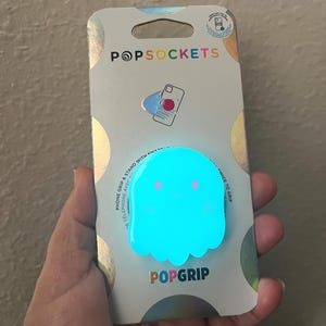 May include: A blue glow-in-the-dark ghost-shaped PopGrip phone grip and stand. The packaging features the PopSockets logo and the text "Phone Grip & Stand with Expandable Telephone Aver... Nice to Grip".