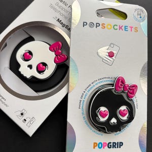 May include: Two PopSockets phone grips and stands in their retail packaging. One features a white skull with pink heart eyes and a pink bow, while the other is a black skull with pink heart eyes and a pink bow. The packaging includes the text PopSockets.