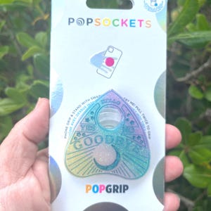 May include: A holographic green, blue, and yellow Ouija board-shaped phone grip with the words "Yes", "No", and "Goodbye" printed on it. The phone grip is on a white background with black polka dots and the words "PopSockets" and "PopGrip" printed on it.