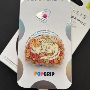 May include: A PopGrip phone accessory featuring a cream-colored dragon sleeping on a pile of orange, yellow, and red autumn leaves. The circular grip is mounted on a white retail card with the text PopSockets and PopGrip printed on it.