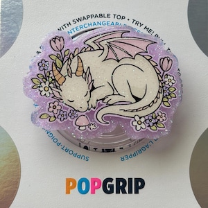 May include: A circular phone grip accessory featuring a glittery lavender resin top with an illustration of a sleeping cream-colored dragon surrounded by small flowers and a mushroom. The base is white with the text POPGRIP printed in black and orange.