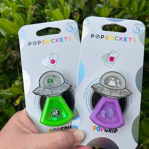 May include: Two PopSockets phone grips with a UFO design. The left grip is green with a glitter alien and the right grip is purple with a glitter ghost.