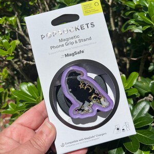 May include: A PopSockets magnetic phone grip and stand for MagSafe. The grip features a purple border with a black center, decorated with gold stars and moons. The packaging is white with the PopSockets logo.