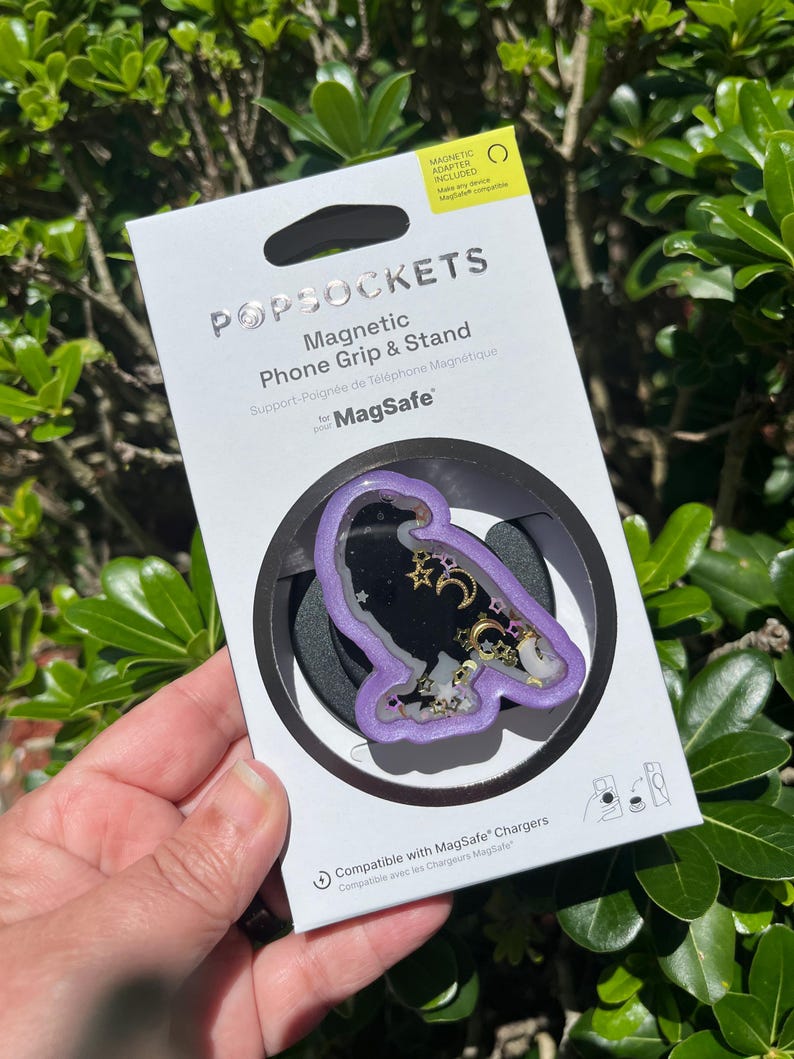 May include: A PopSockets magnetic phone grip and stand for MagSafe. The grip features a purple border with a black center, decorated with gold stars and moons. The packaging is white with the PopSockets logo.