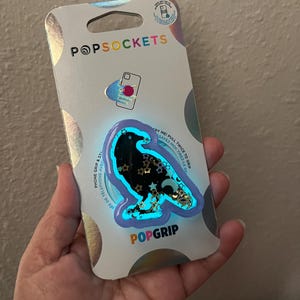 May include: A PopSockets PopGrip phone accessory featuring a black raven design with gold stars and a blue outline. The packaging is silver with the PopSockets logo and the words "Phone Grip & Stand" and "Sayez-Moi Tirez-Le Del".