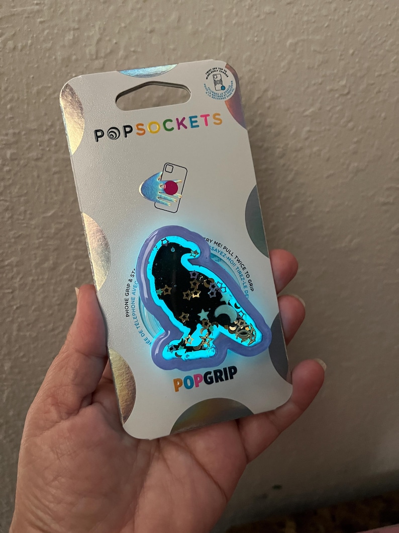 May include: A PopSockets PopGrip phone accessory featuring a black raven design with gold stars and a blue outline. The packaging is silver with the PopSockets logo and the words "Phone Grip & Stand" and "Sayez-Moi Tirez-Le Del".