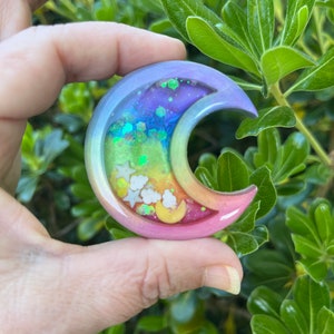 May include: A rainbow crescent moon resin charm with glitter and white stars. The charm is held in a hand against a green leafy background.