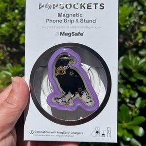 May include: A PopSockets Magnetic Phone Grip & Stand for MagSafe. The grip features a black owl design with gold stars and moons, encased in a clear, purple-edged frame. The packaging includes the text "MAGNETIC ADAPTER INCLUDED".