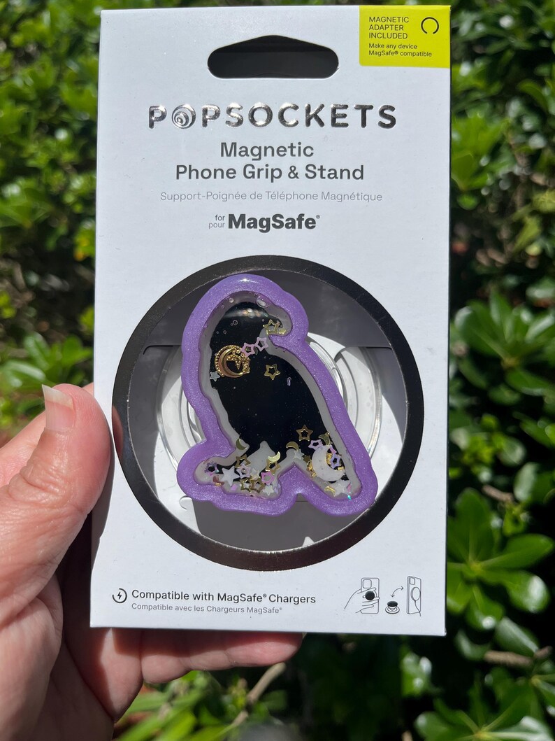 May include: A PopSockets Magnetic Phone Grip & Stand for MagSafe. The grip features a black owl design with gold stars and moons, encased in a clear, purple-edged frame. The packaging includes the text "MAGNETIC ADAPTER INCLUDED".