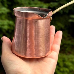 Peut inclure: Une petite cafetière en cuivre avec une poignée en laiton. La cafetière mesure environ 12,5 cm de haut et a un fond arrondi.