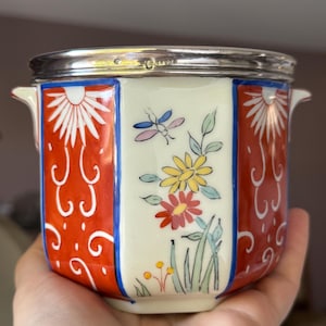 Puede incluir: Maceta octogonal de porcelana con borde plateado. La maceta tiene un fondo blanco con diseños florales rojos, azules y amarillos y una libélula.
