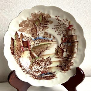Könnte beinhalten: Eine weiße Keramik-Untertasse mit einem braunen und grünen Blumenmuster. Die Untertasse zeigt eine Szene mit einer Kutsche, die ein Landgasthaus verlässt. Der Text "Leaving the Inn" ist auf der Untertasse aufgedruckt.