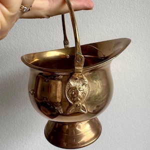 Puede incluir: Un pequeño cubo de metal dorado con asa. El cubo tiene un diseño decorativo en el lateral y una base redonda.