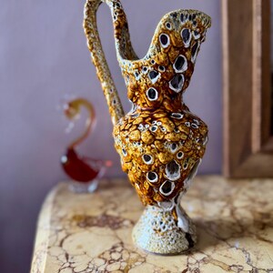May include: A textured, ceramic pitcher with a handle. The pitcher is primarily brown and tan with white accents and a unique, porous design. The pitcher is on a marble surface, with a glass swan in the background.