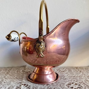 Könnte beinhalten: Eine kupferfarbene Zuckerdose mit einem Messinggriff und dekorativen Löwenkopf-Akzenten. Die Dose hat einen Ausguss und einen kleinen Griff mit einem weiß-schwarzen Tupfenmuster. Sie steht auf einem Sockel.