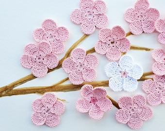 May include: A collection of pink and white crocheted flowers, resembling cherry blossoms, arranged on a brown branch painted on a white background.