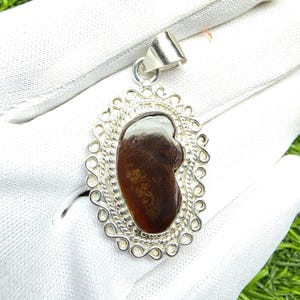 May include: A pendant featuring an irregular, dark brown, translucent stone set in a polished silver frame. The setting includes a beaded inner border and an outer decorative border of swirling silver wire, topped with a triangular silver bail.