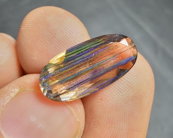 Puede incluir: Una piedra preciosa de corte ovalado con una mezcla de colores, incluyendo naranja, verde y azul. La piedra preciosa tiene un patrón rayado y se sostiene entre los dedos. La gema está facetada, reflejando la luz.
