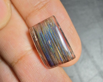 Puede incluir: Un cabujón de piedra preciosa rectangular y pulido con una base clara y transparente, que presenta densas inclusiones paralelas en forma de aguja en tonos azul iridiscente, dorado y cobre, creando un efecto metálico multicolor.