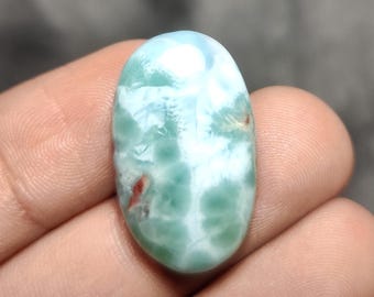 May include: An oval-shaped, polished gemstone with a light blue and white pattern, featuring areas of green and small red inclusions. The stone is held in a hand, showcasing its natural beauty and unique color variations. This gemstone is ideal for jewelry making.