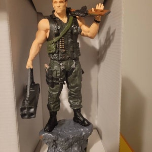 May include: A green and black action figure of a soldier holding a gun and a rocket launcher. The figure is standing on a grey rock base with the word "COMMANDO" written on it.