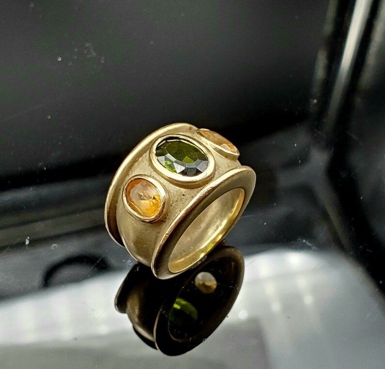 May include: A gold ring featuring three oval-shaped gemstones. The central stone is green, with two yellow stones flanking it.