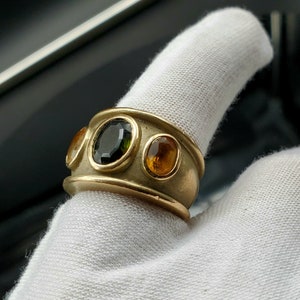 May include: A gold ring featuring three gemstones. The central stone is a dark green oval, and the two flanking stones are a lighter orange oval.