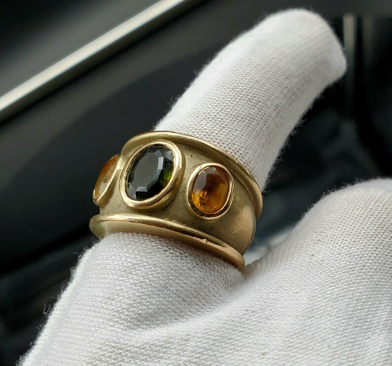 May include: A gold ring featuring three gemstones. The central stone is a dark green oval, and the two flanking stones are a lighter orange oval.