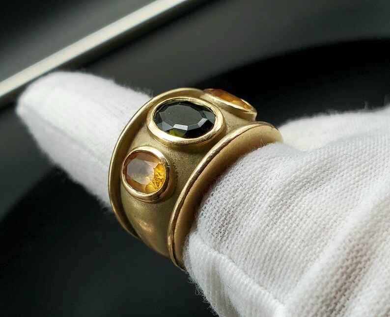 May include: A gold ring featuring three oval-shaped gemstones. The central stone is black, and the two outer stones are yellow.